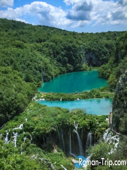 A stunning panoramic view of the Plitvice Waterfalls, capturing the full grandeur of this iconic natural wonder. The powerful cascade of water plunges into the vibrant turquoise pools below, surrounded by lush greenery in the heart of Plitvice Lakes National Park.