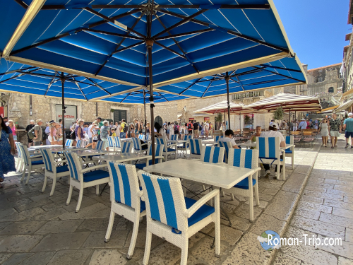 ドゥブロブニク旧市街のカフェテラス。青いパラソルとストライプの椅子が並び、観光客でにぎわう夏の街角。/ A café terrace in Dubrovnik’s Old Town with blue parasols and striped chairs, bustling with tourists on a sunny summer day.