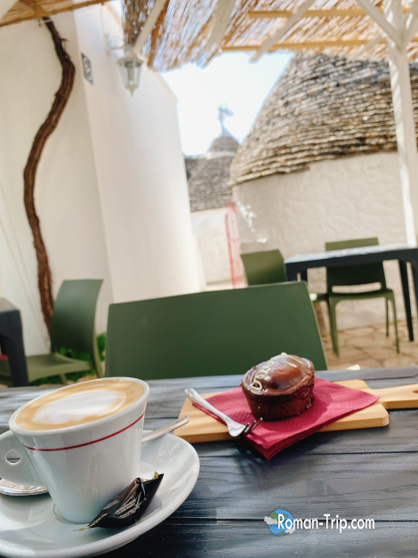 アルベロベッロのカフェで、とんがり屋根を眺めながら味わうカプチーノとケーキ。旅の癒しのひととき。/ Enjoying cappuccino and cake at a café in Alberobello, with a view of the charming trulli rooftops.