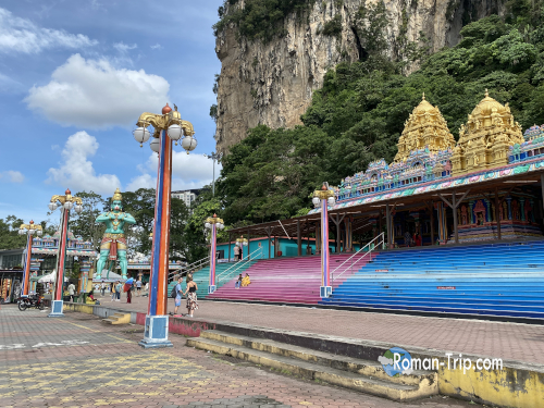 バトゥ洞窟の駅を降りたときの景色。そびえ立つ崖とカラフルな建物が目を引く。/ View from Batu Caves station, featuring towering cliffs and colorful buildings.