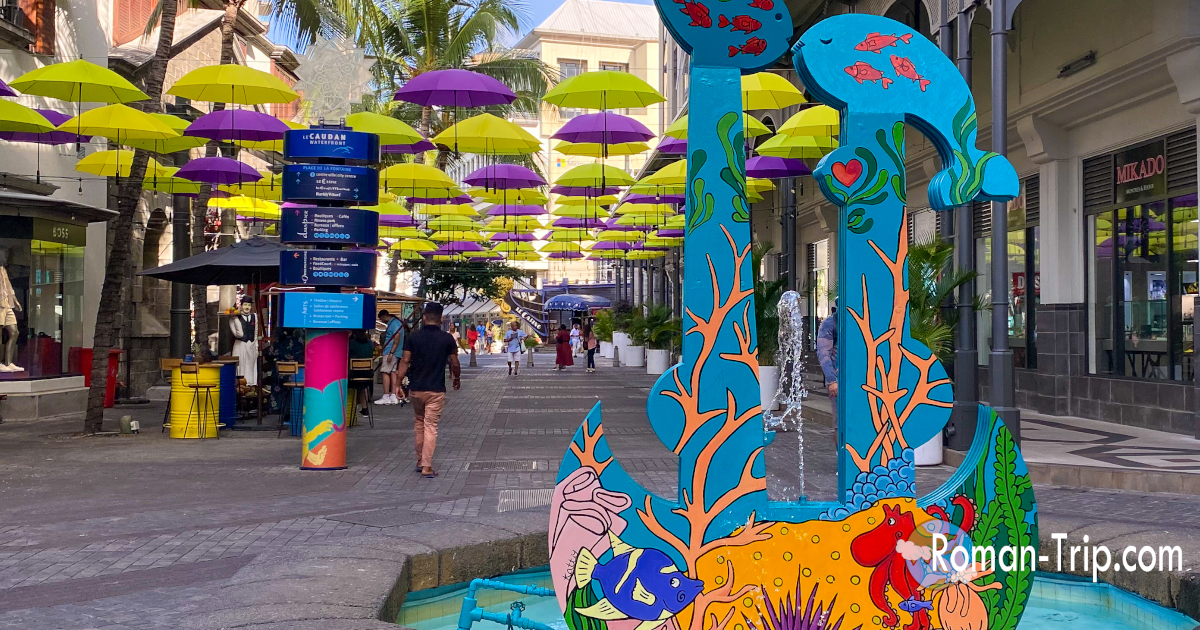 モーリシャスの首都ポートルイス、コーダンウォーターフロントのアンブレラストリート。黄色と紫の傘が並び、カラフルな錨のアートが旅気分を盛り上げる。/ Umbrella Street at the Caudan Waterfront in Port Louis, Mauritius — yellow and purple umbrellas and a colorful anchor sculpture creating a vibrant travel atmosphere.