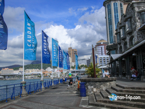 モーリシャスの首都ポートルイス、コーダンウォーターフロントの海沿いプロムナード。青い旗が並び、穏やかな海風が吹き抜ける。/ The seaside promenade at the Caudan Waterfront in Port Louis, Mauritius, lined with blue flags fluttering in the gentle ocean breeze.