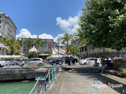 モーリシャスの首都ポートルイス、コーダンウォーターフロントの通り沿いの風景。ヤシの木と海風が心地よいリゾートエリア。/ Street view of the Caudan Waterfront in Port Louis, Mauritius, lined with palm trees and a relaxing seaside atmosphere.