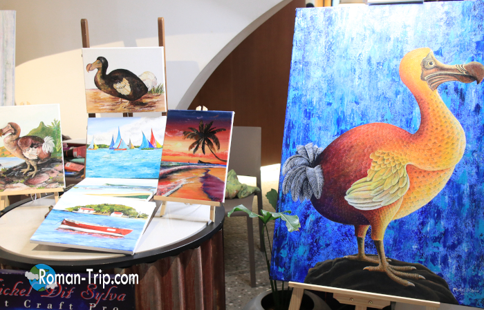 モーリシャスのクラフトマーケットで見つけたドードーの絵画展示。青い背景に描かれた大きなドードーと、南国の風景画が並ぶ。/ Dodo paintings displayed at the Craft Market in Port Louis, Mauritius — featuring a large blue-background dodo and colorful tropical landscapes.