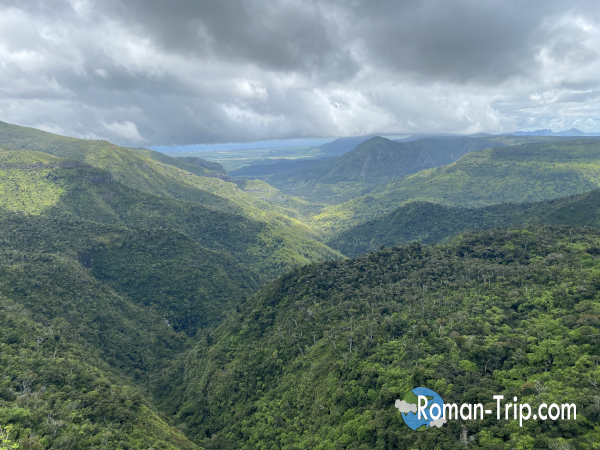 モーリシャスのゴルジュ展望台から望む大渓谷のパノラマ。深い緑に覆われた山々と、雲の切れ間から差し込む光が壮大な自然を際立たせる。/ Panoramic view from the Gorges viewpoint in Mauritius, overlooking vast green valleys and mountains illuminated by sunlight breaking through the clouds.