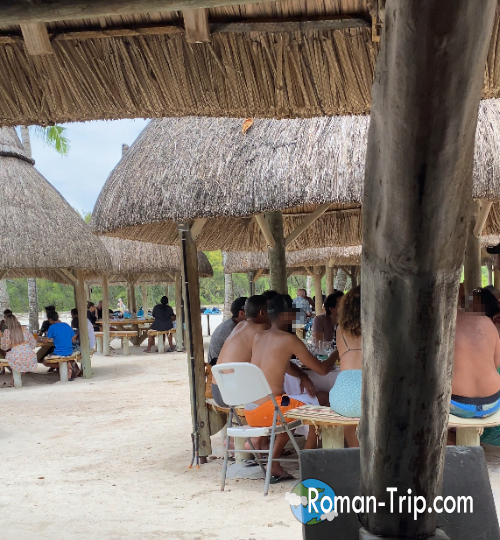 モーリシャスのイル・オ・セルフでビュッフェを楽しみながら談笑する観光客たち。ヤシ葺き屋根の下でゆったり過ごす南国らしい時間。/ Tourists enjoying a buffet lunch and chatting under thatched huts at Île aux Cerfs, Mauritius — a relaxed tropical dining atmosphere.