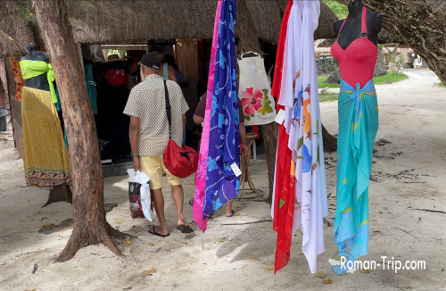 モーリシャスのイル・オ・セルフ島にあるお土産屋の外観。ビーチドレスやパレオが吊るされ、砂浜沿いで買い物を楽しむ観光客の姿が見える。/ Souvenir shop at Île aux Cerfs, Mauritius, displaying colorful beach dresses and pareos outside, with visitors browsing in a sandy tropical setting.