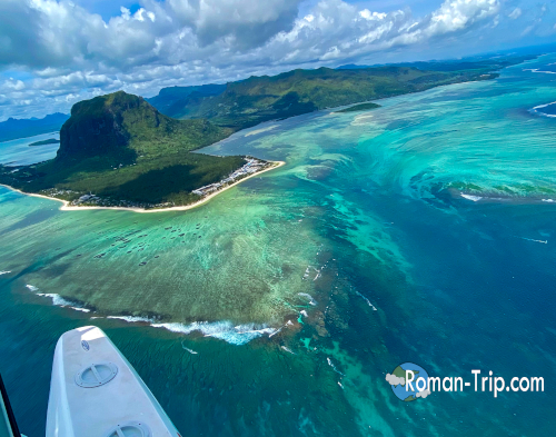 モーリシャス観光のハイライト、海の滝とルモーン山。上空から望むと、海底の砂と潮流が生み出す幻想的な「海の滝」と、世界遺産ルモーン山が並ぶ壮大な景色が広がる。 / A highlight of Mauritius sightseeing: the underwater waterfall and Le Morne Brabant. Seen from above, the illusion of a cascading waterfall beneath the sea appears alongside the iconic Le Morne mountain, creating a truly spectacular view.