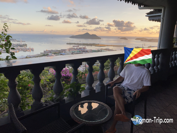 セーシェル・ヴィクトリアのBlue Hill Guest Houseのテラスで、夕景を背景に椅子に腰掛け脚を組みながら自分を写した一枚。港と海に沈む夕日の時間を記録した瞬間。 / A self-photo taken on the terrace of Blue Hill Guest House in Victoria, Seychelles, sitting on a chair with legs crossed while capturing the sunset view over the harbor and sea.