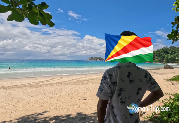 セーシェルのグランド・アンス・ビーチで記念撮影をする様子。青い海と白い砂浜を背景にした旅の思い出の一枚。 / A memorable photo taken at Grand Anse Beach in Seychelles, with the blue ocean and white sandy beach as the backdrop.