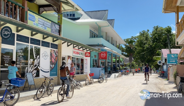 セーシェル・ラディーグ島の唯一の通り。自転車で移動する人々やカラフルな建物が並ぶ、島らしいのどかな街並み。 / The main street of La Digue Island in Seychelles, where people travel by bicycle along a quiet road lined with colorful buildings and small shops.
