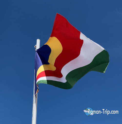 セーシェルのラディーグ港になびくセーシェル国旗。青・黄・赤・白・緑の鮮やかな旗が南国の港で風に揺れる。 / The Seychelles national flag waving at La Digue Port, its bright blue, yellow, red, white, and green colors fluttering in the tropical harbor breeze.