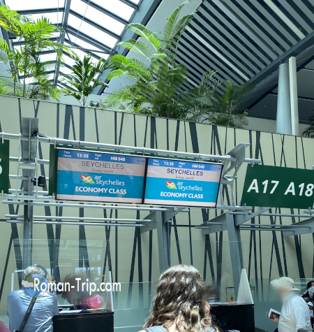 セーシェル行きフライトのチェックインカウンター。空港の大きな窓から明るい日差しが差し込み、南国らしい開放的な雰囲気に包まれている。 / Check-in counter for a flight to Seychelles, where bright tropical sunlight streams through the airport windows, creating a warm and relaxed island atmosphere.